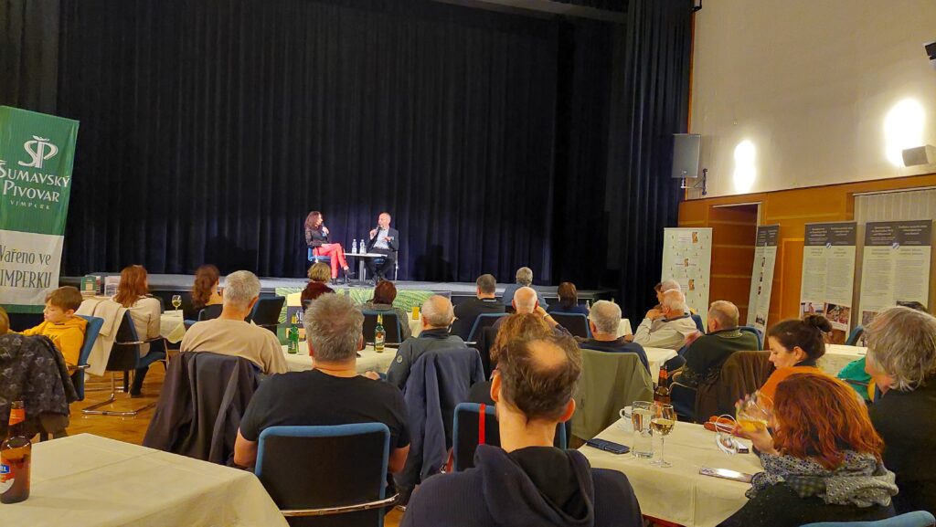 Diskuse na literárním festivalu "Šumava-Litera". Diváci sedí u stolů a dívají se na pódium. Diskutují Václava Jandečková a Martin Sichinger. 